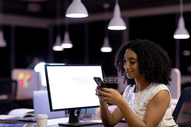 Вид збоку молодої змішаної професійної жінки, яка працює пізно в сучасному офісі, сидить за столом за допомогою смартфона — стокове фото