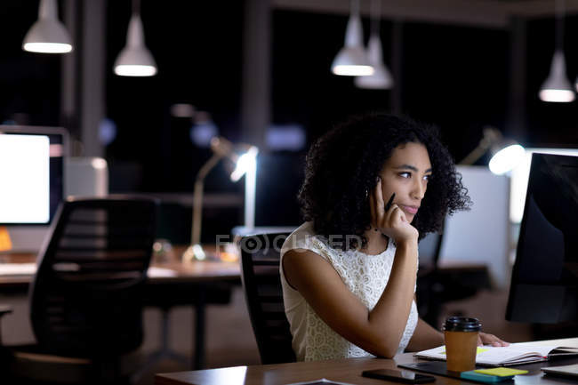 Vista lateral de una joven mujer profesional de raza mixta que trabaja hasta tarde en una oficina moderna, sentada en un escritorio usando una computadora de escritorio - foto de stock