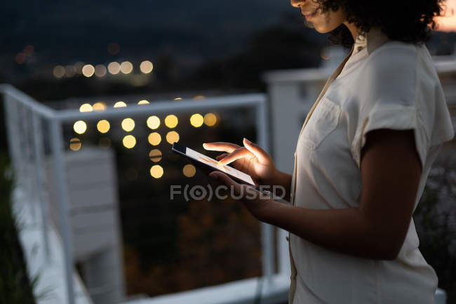 Бічний вид молодої професійної жінки змішаної раси, яка працює пізно в сучасному офісі, стоїть на терасі даху за допомогою планшетного комп'ютера. — стокове фото