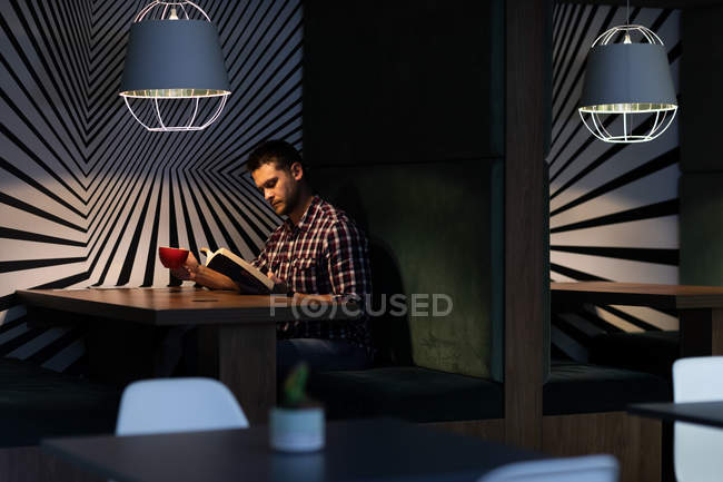 Вид сбоку на молодого кавказского профессионала, работающего допоздна в современном офисе, сидящего за столом, читающего книгу и держащего чашку — стоковое фото