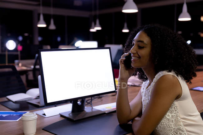 Vista lateral de una joven profesional de raza mixta que trabaja hasta tarde en una oficina moderna, sentada en un escritorio hablando en un teléfono inteligente - foto de stock