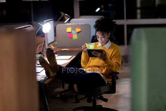 Вид спереди на молодую кавказскую профессиональную расистку, работающую допоздна в современном офисе, сидящую за столом с поднятыми ногами за планшетным компьютером и держащую чашку — стоковое фото