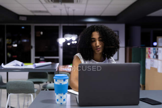 Вид спереду молода змішана професійна жінка, що працює пізно в сучасному офісі, сидить за столом з кавою на винос за допомогою ноутбука — стокове фото