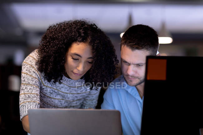 Vista frontal close-up de um jovem homem profissional caucasiano e mulher de raça mista trabalhando tarde em um escritório moderno em uma mesa usando um computador portátil juntos — Fotografia de Stock