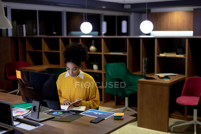 Frontansicht einer jungen Frau mit gemischter Rasse, die spät in einem modernen Büro arbeitet, am Schreibtisch sitzt und ein Buch liest — Stockfoto