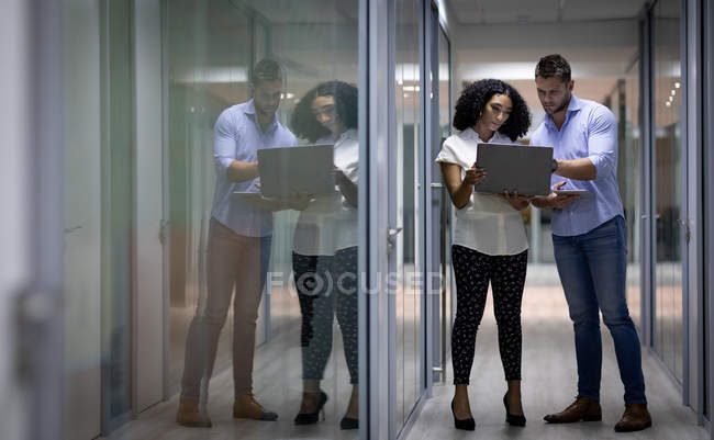 Vista frontal de un joven hombre profesional caucásico y una mujer de raza mixta que trabaja hasta tarde en una oficina moderna, de pie en el pasillo utilizando una computadora portátil, reflejada en una pared de vidrio - foto de stock