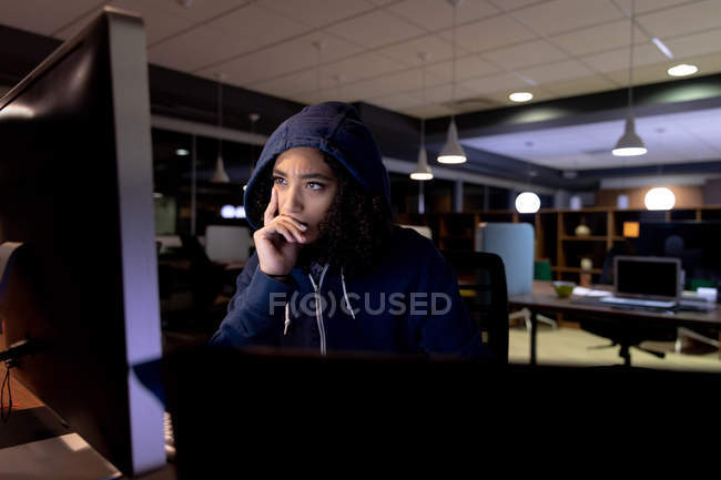 Vista frontal de una joven mujer de raza mixta profesional caucásica con una sudadera con capucha que trabaja hasta tarde en una oficina moderna, sentada en un escritorio apoyado y mirando fijamente a un monitor de computadora de escritorio - foto de stock