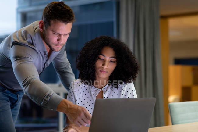 Vista frontal de um jovem homem profissional caucasiano e mulher de raça mista trabalhando tarde em um escritório moderno em uma mesa usando um computador portátil, o homem de pé e apontando para a tela — Fotografia de Stock