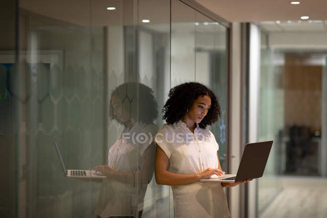 Vista frontal de una joven profesional de raza mixta que trabaja hasta tarde en una oficina moderna, de pie en el pasillo usando una computadora portátil, reflejada en una pared de vidrio - foto de stock