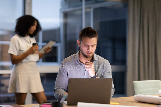 Передній погляд на молодого кавказького професіонала і змішаної расової жінки, що працює в сучасному офісі, чоловіка, який сидить за столом з ноутбуком, а жінка стоїть на задньому плані, тримаючи табличку. — стокове фото