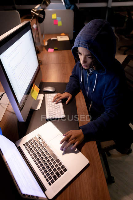 Blick aus der Vogelperspektive auf eine junge Frau, die einen Kapuzenpullover trägt und spät in einem modernen Büro an einem Schreibtisch sitzt, Laptop und Desktop-Computer benutzt — Stockfoto