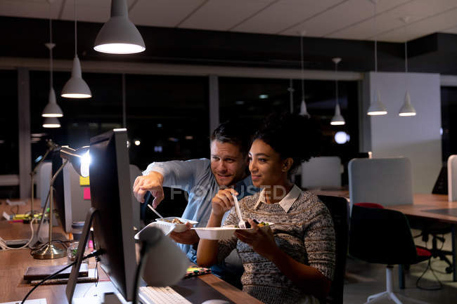 Vue de face d'un jeune homme professionnel caucasien et d'une femme de race mixte travaillant tard dans un bureau moderne, debout à un bureau à l'aide d'un ordinateur portable — Photo de stock