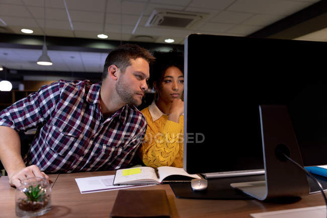Frontansicht eines jungen kaukasischen Berufsmannes und einer Frau mit gemischter Rasse, die spät in einem modernen Büro arbeiten und am Schreibtisch mit einem Desktop-Computer sitzen — Stockfoto