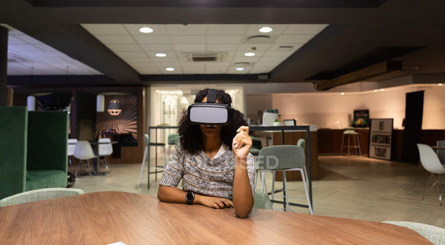 Передній вигляд молодої змішаної професійної жінки, яка працює пізно в сучасному офісі, сидить за столом у гарнітурі VR — стокове фото