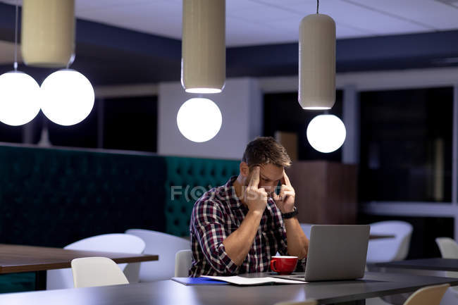 Vista frontale di un giovane professionista caucasico che lavora fino a tardi in un ufficio moderno, seduto a una scrivania che tiene la testa e fissa un computer portatile, con una tazza di caffè di fronte a lui — Foto stock