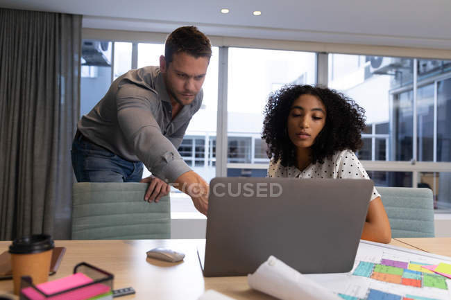 Вид спереди на молодого кавказского профессионального мужчину и женщину смешанной расы, работающую в современном офисе за столом, используя ноутбук, мужчину, стоящего и указывающего на экран — стоковое фото