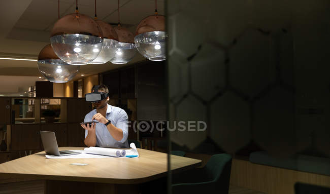 Вид молодого кавказца - профессионала, работающего поздно вечером в современном офисе, сидящего за столом с ноутбуком, планшетным компьютером и носящего гарнитуру. — стоковое фото