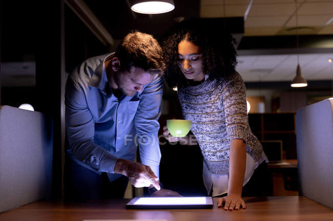 Вид спереди на молодого кавказского профессионала и женщину смешанной расы, работающую допоздна в современном офисе, стоящую за столом, смотрящую вместе на планшетный компьютер, мужчину, указывающего на экран, и женщину, держащую чашку — стоковое фото