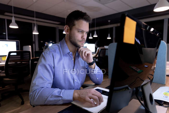 Vista frontal de um jovem homem profissional caucasiano trabalhando até tarde em um escritório moderno, sentado em uma mesa usando um computador desktop — Fotografia de Stock