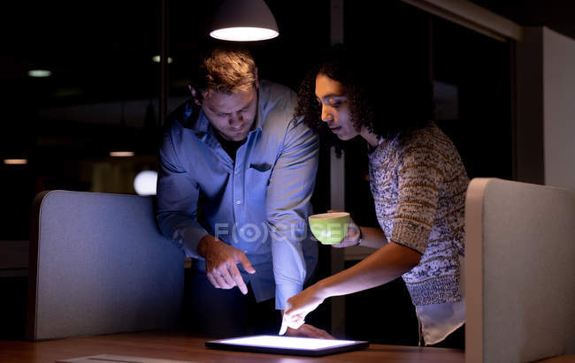 Вид спереди на молодого кавказского профессионального мужчину и женщину смешанной расы, работающую допоздна в современном офисе, стоящую за столом, смотрящую на планшетный компьютер вместе, женщину, трогающую экран и держащую чашку — стоковое фото