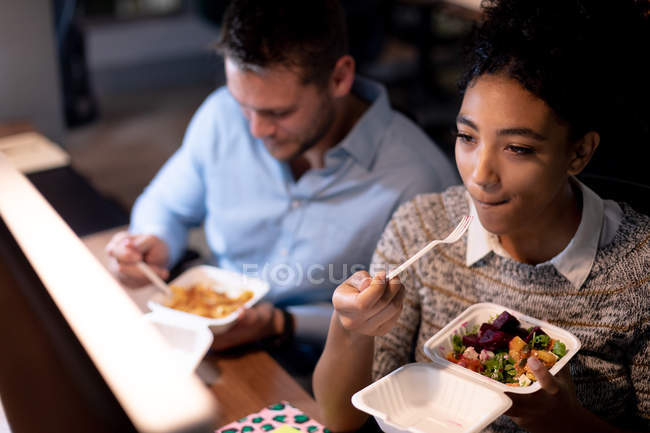 Vue en angle élevé d'un jeune homme professionnel caucasien et d'une femme de race mixte travaillant tard dans un bureau moderne, assis à un bureau mangeant de la nourriture à emporter ensemble, la femme regardant un écran d'ordinateur pendant qu'elle mange — Photo de stock