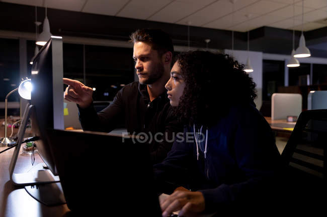 Seitenansicht eines jungen kaukasischen Berufsmannes und einer Frau mit gemischter Rasse, die spät in einem modernen Büro arbeiten, am Schreibtisch einen Laptop benutzen und gemeinsam auf einen Desktop-Computermonitor blicken, wobei der Mann auf den Bildschirm zeigt. — Stockfoto