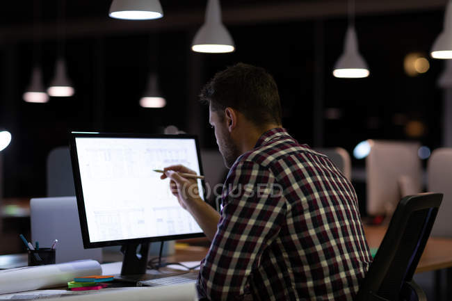 Visão traseira de um jovem homem profissional caucasiano trabalhando até tarde em um escritório moderno, sentado em uma mesa olhando para um monitor de computador e apontando com um lápis — Fotografia de Stock