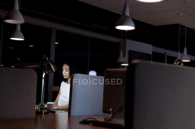 Vista lateral de uma jovem mulher profissional de raça mista trabalhando tarde em um escritório moderno, sentado em uma mesa com um café takeaway usando um computador desktop — Fotografia de Stock