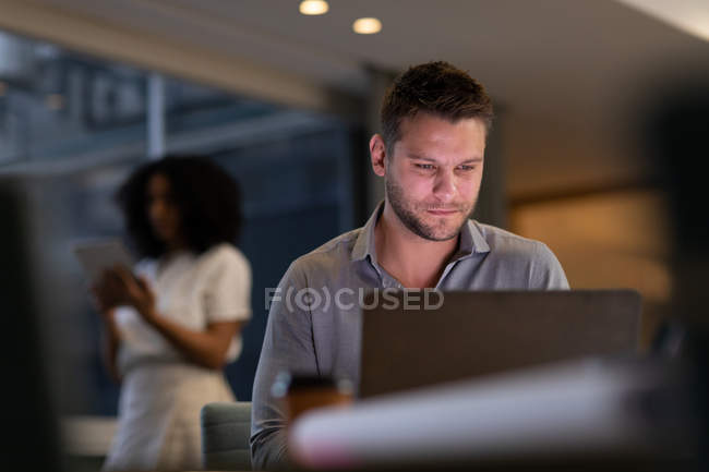 Вид спереди на молодого кавказского профессионального мужчину и женщину смешанной расы, работающую допоздна в современном офисе, мужчину, сидящего за столом с помощью ноутбука, и женщину, стоящую на заднем плане с помощью планшета — стоковое фото