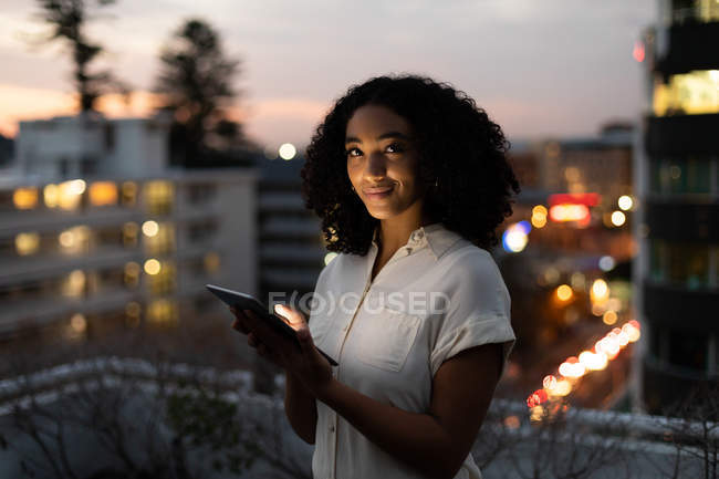 Retrato de una joven profesional de raza mixta que trabaja hasta tarde en una oficina moderna, de pie en la terraza de la azotea usando una tableta, sonriendo a la cámara - foto de stock