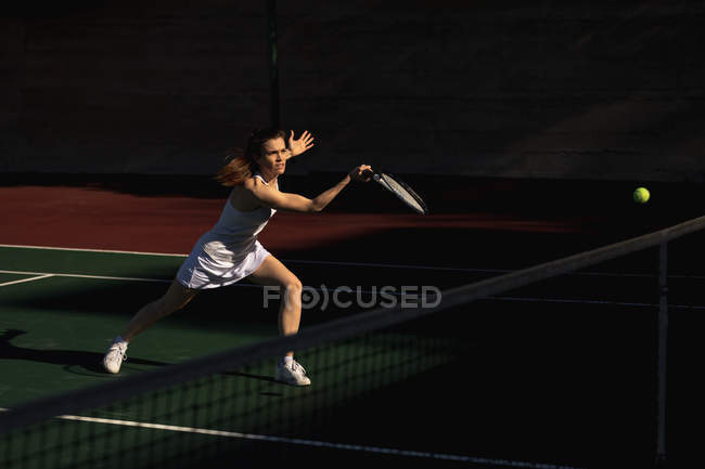 Vue latérale d'une jeune femme caucasienne et d'un homme jouant au tennis par une journée ensoleillée, femme courant jusqu'au ballon — Photo de stock