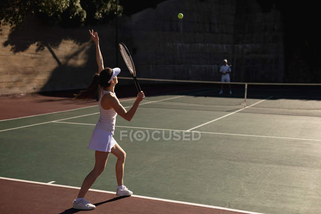 Vista lateral de una joven mujer caucásica y un hombre jugando tenis en un día soleado, mujer golpeando la pelota - foto de stock