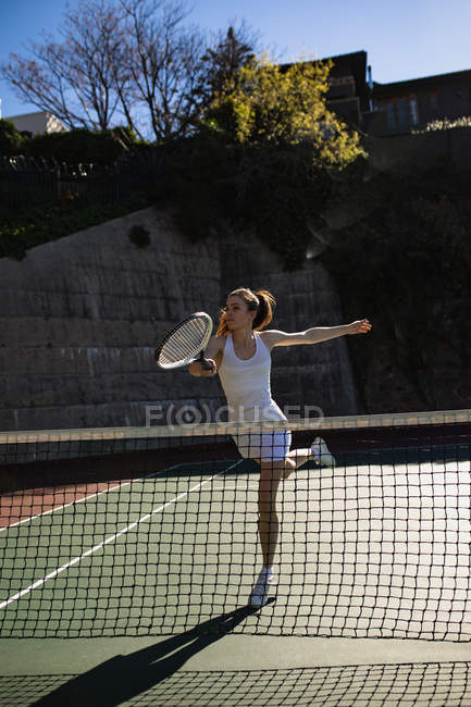 Vista frontal de una joven mujer caucásica jugando tenis en un día soleado, corriendo a golpear una pelota con una pared detrás de ella - foto de stock