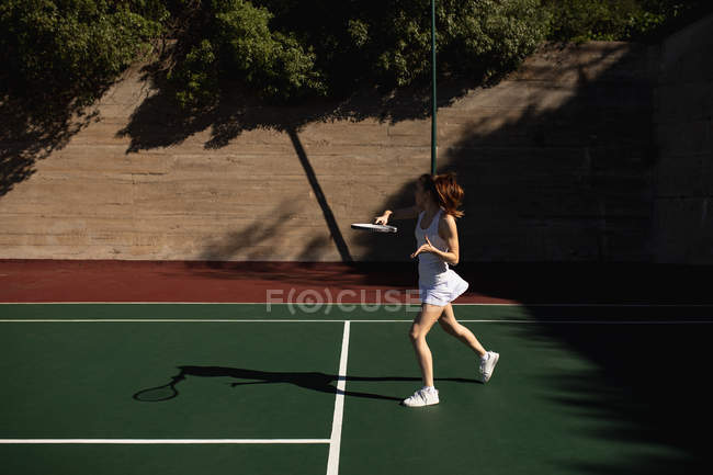 Vista lateral de uma jovem caucasiana jogando tênis em um dia ensolarado, batendo uma bola com uma parede atrás dela — Fotografia de Stock