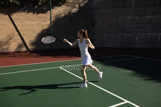 Vista lateral de una joven mujer caucásica jugando al tenis en un día soleado, corriendo hasta la pelota con una pared detrás de ella - foto de stock