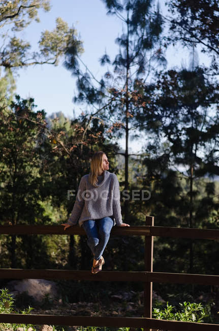 Comprimento total de caminhante feminino pensativo sentado em cerca de madeira contra árvores — Fotografia de Stock