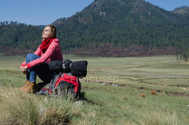 Volle Länge einer Wanderin, die aufschaut, während sie auf einem Felsen gegen den Berg sitzt — Stockfoto