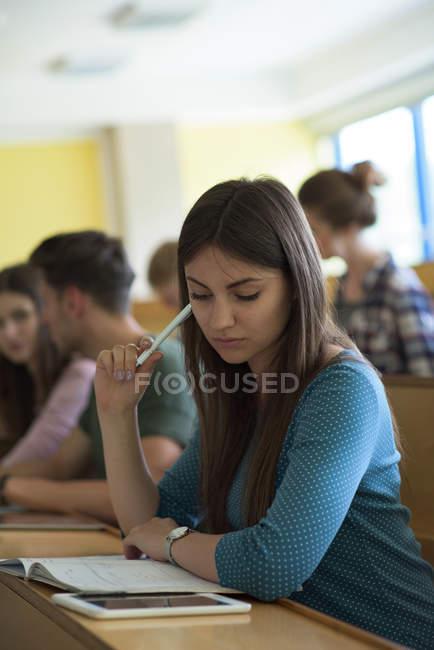 Young female using tablet computer while studying at desk with classmates at desk in classroom — Stock Photo