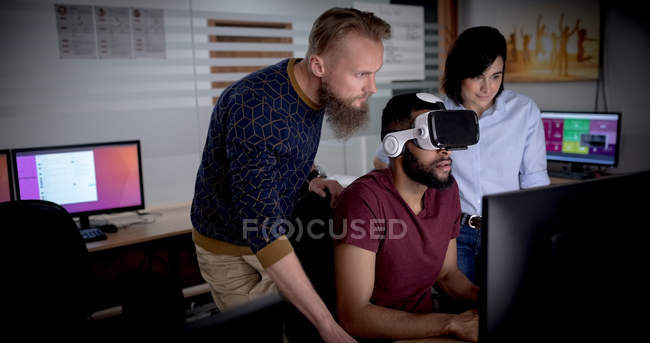 Коллеги, использующие гарнитуру виртуальной реальности во время работы на компьютере в офисе — стоковое фото