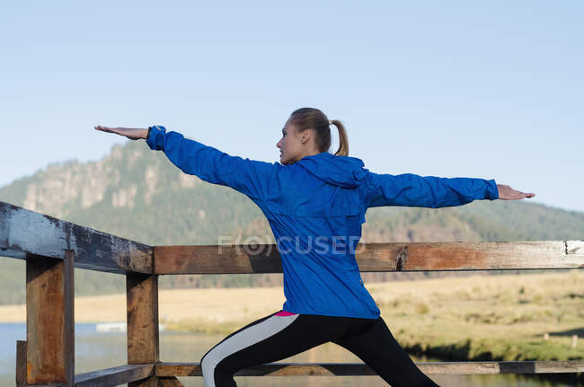 Rear view of female athlete practicing Warrior 2 pose while exercising on pier — Stock Photo