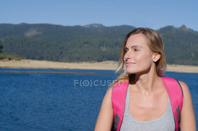 Primer plano de senderista sonriente mirando hacia otro lado mientras está de pie contra el lago - foto de stock