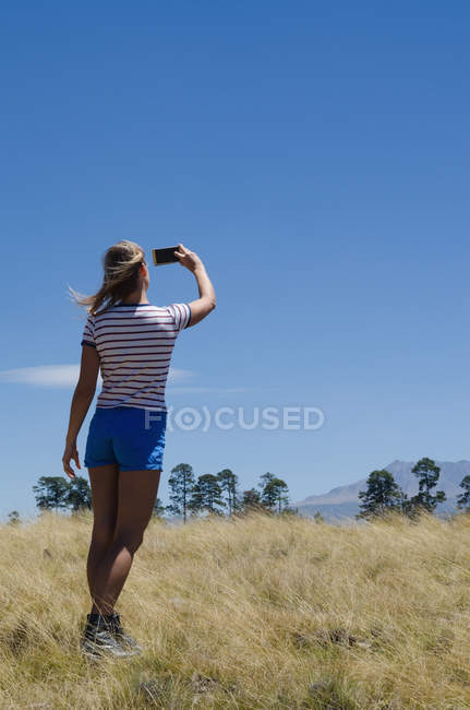 Вид ззаду на жіночого пішохода, що приймає селфі, стоячи на полі — стокове фото
