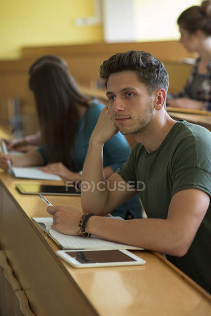 Estudante do sexo masculino pensativo com colegas na mesa em sala de aula — Fotografia de Stock