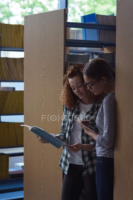 Female students reading book while standing by shelf in library — Stock Photo