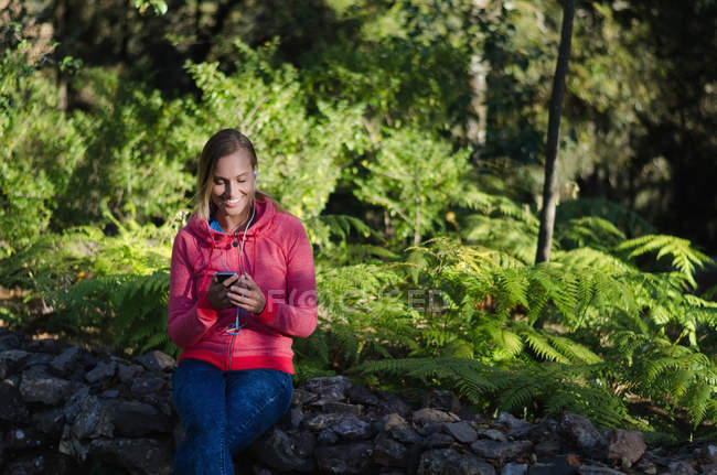 Caminhante sorridente feminino usando telefone enquanto sentado na rocha na floresta — Fotografia de Stock