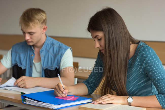 Estudante feminina escrevendo em nota adesiva enquanto estudava na mesa em sala de aula — Fotografia de Stock
