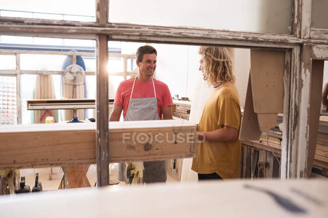 Two Caucasian male surfboard makers standing in their studio, preparing for work, discussing the project, with ready surfboards in the background. — Stock Photo