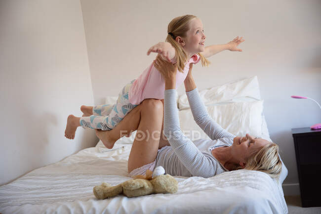 Vista lateral de una mujer caucásica disfrutando de tiempo en familia con su hija en casa juntos, acostada en una cama en su dormitorio sonriendo y levantándola por encima de ella - foto de stock