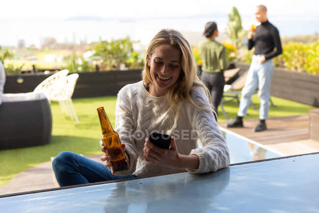 Вид спереди на белую женщину, болтающуюся на террасе на крыше в солнечный день, использующую смартфон и держащую бутылку пива, улыбающуюся, с людьми, говорящими на заднем плане — стоковое фото