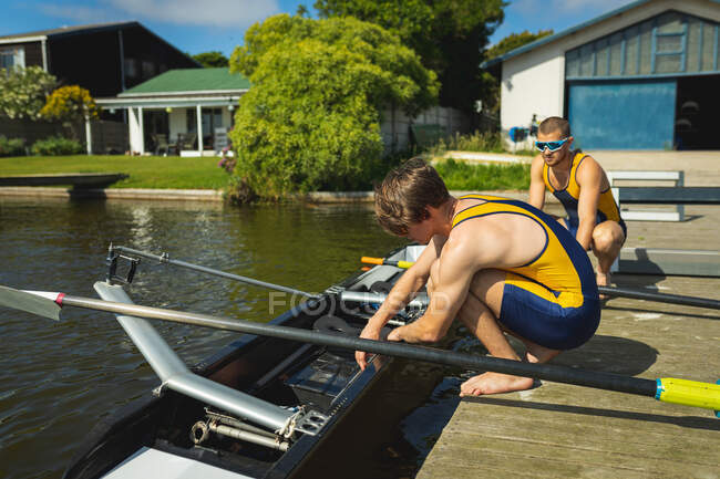Side view of a rowing team of two Caucasian men putting rowing boat into the water before rowing, crouching on a jetty on the river on a sunny day — Stock Photo